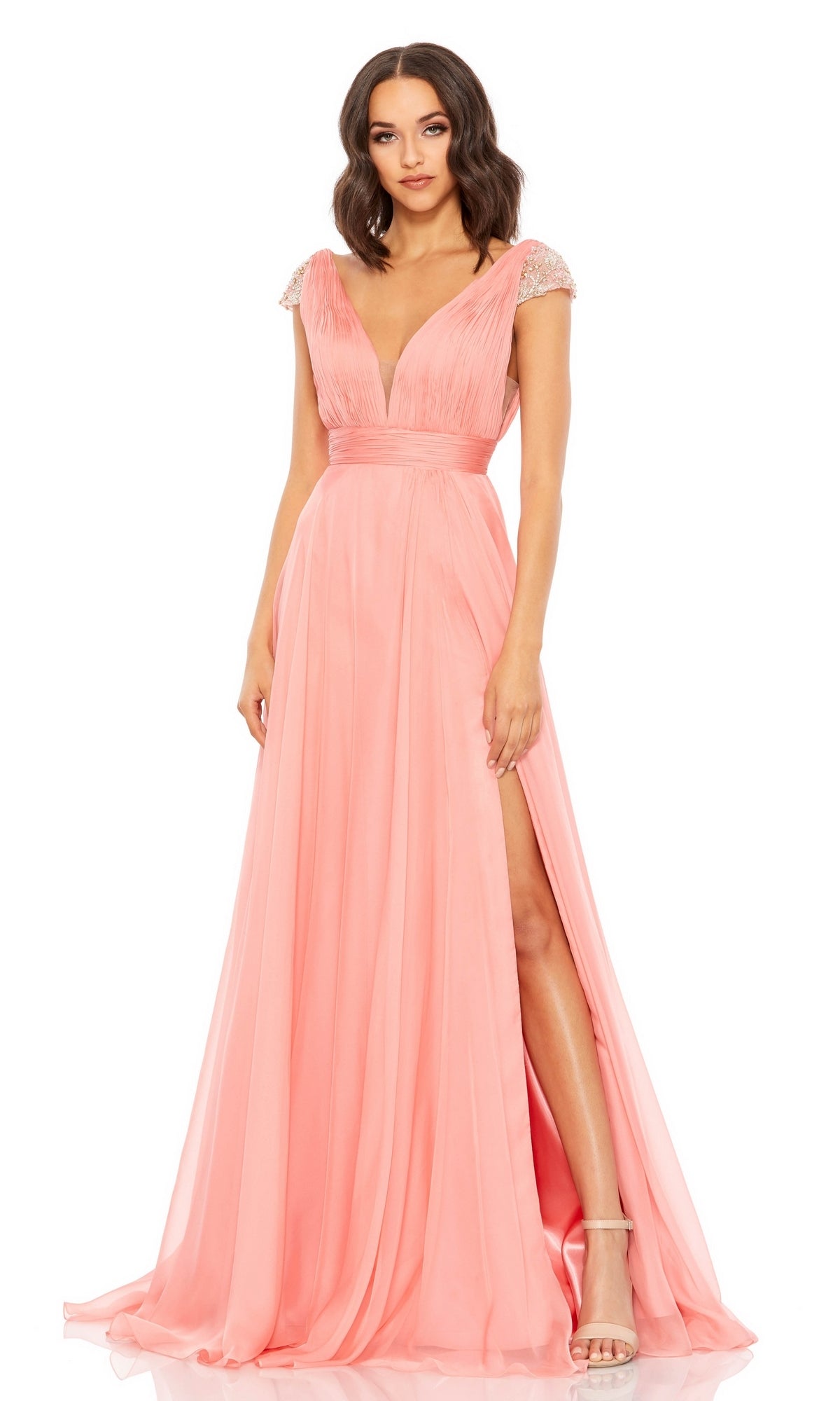 A woman wears the Coral Pink Cap Sleeve Long Prom Gown 67811, featuring beaded cap sleeves, a deep V-neckline, and a thigh-high slit, paired with nude high-heeled sandals.